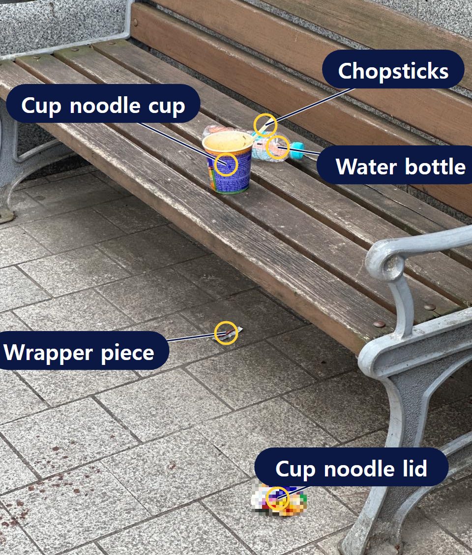 Photo of a cup noodle container, a water bottle and wooden chopsticks on a bench, with the cup noodle lid and a small piece of packaging on the ground