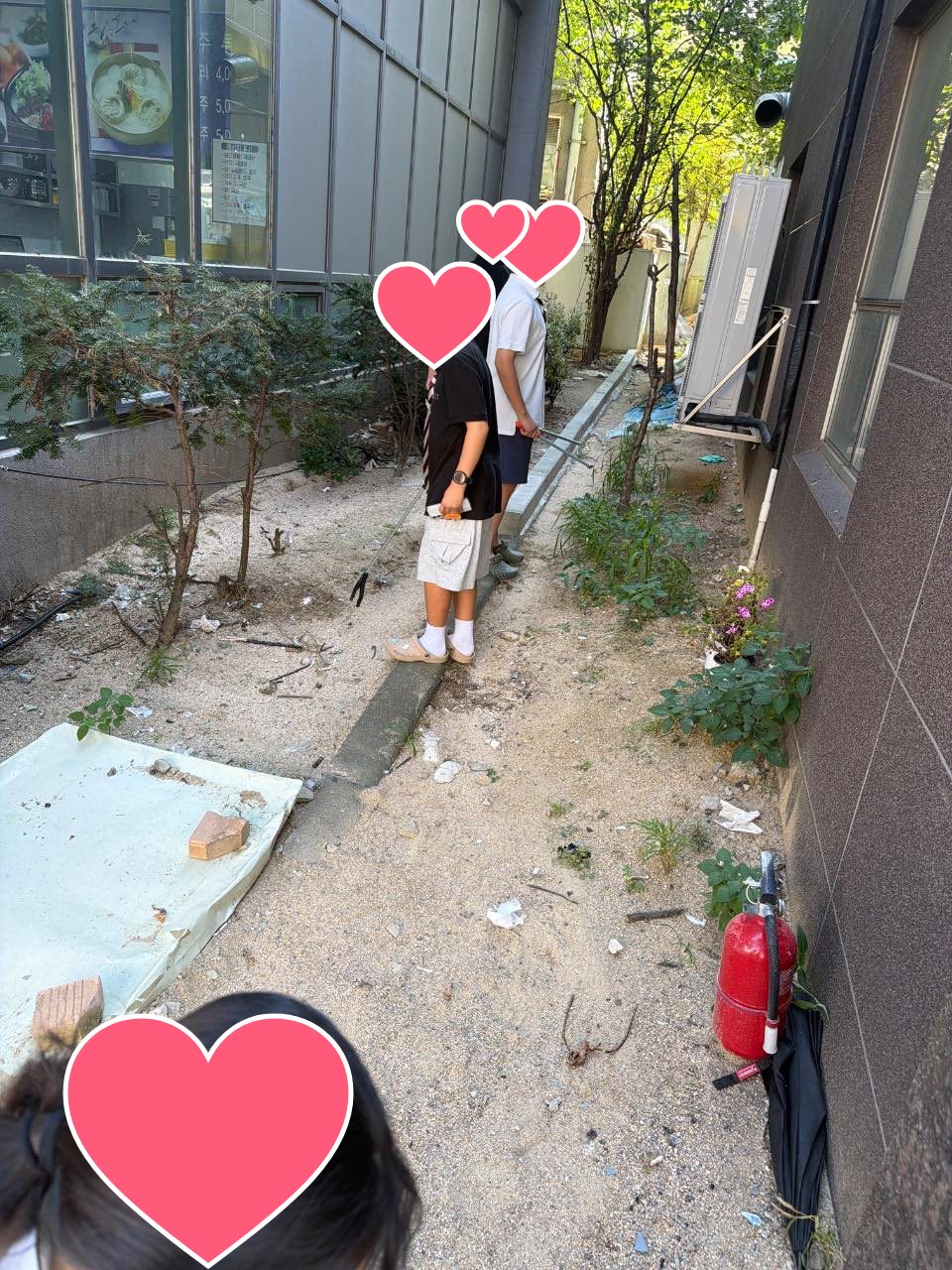 Students picking up trash with friends in a narrow alley beside the school building (faces covered with hearts)